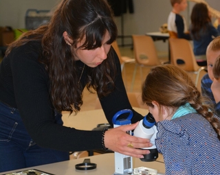 Vue d'un atelier scientifique avec un enfant qui regarde dans une loupe binoculaire, aidé par une médiatrice scientifique