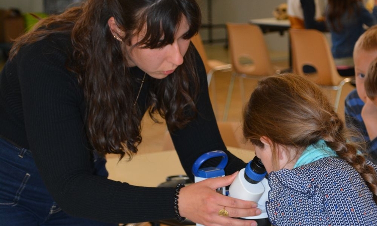Vue d'un atelier scientifique avec un enfant qui regarde dans une loupe binoculaire, aidé par une médiatrice scientifique